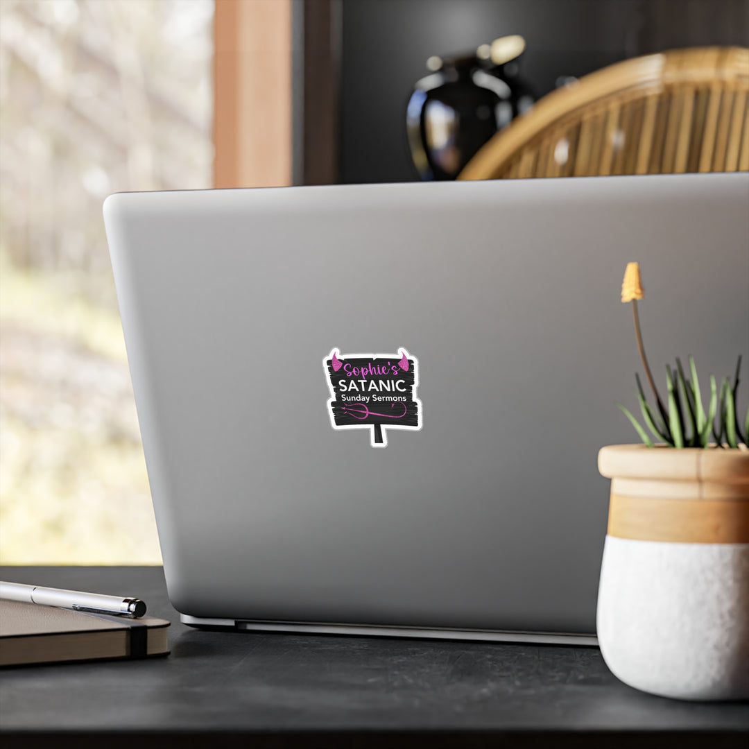 A sticker with text "Sophie's Satanic Sunday Sermons" placed on a laptop which is on a desk, next to a potted plant.