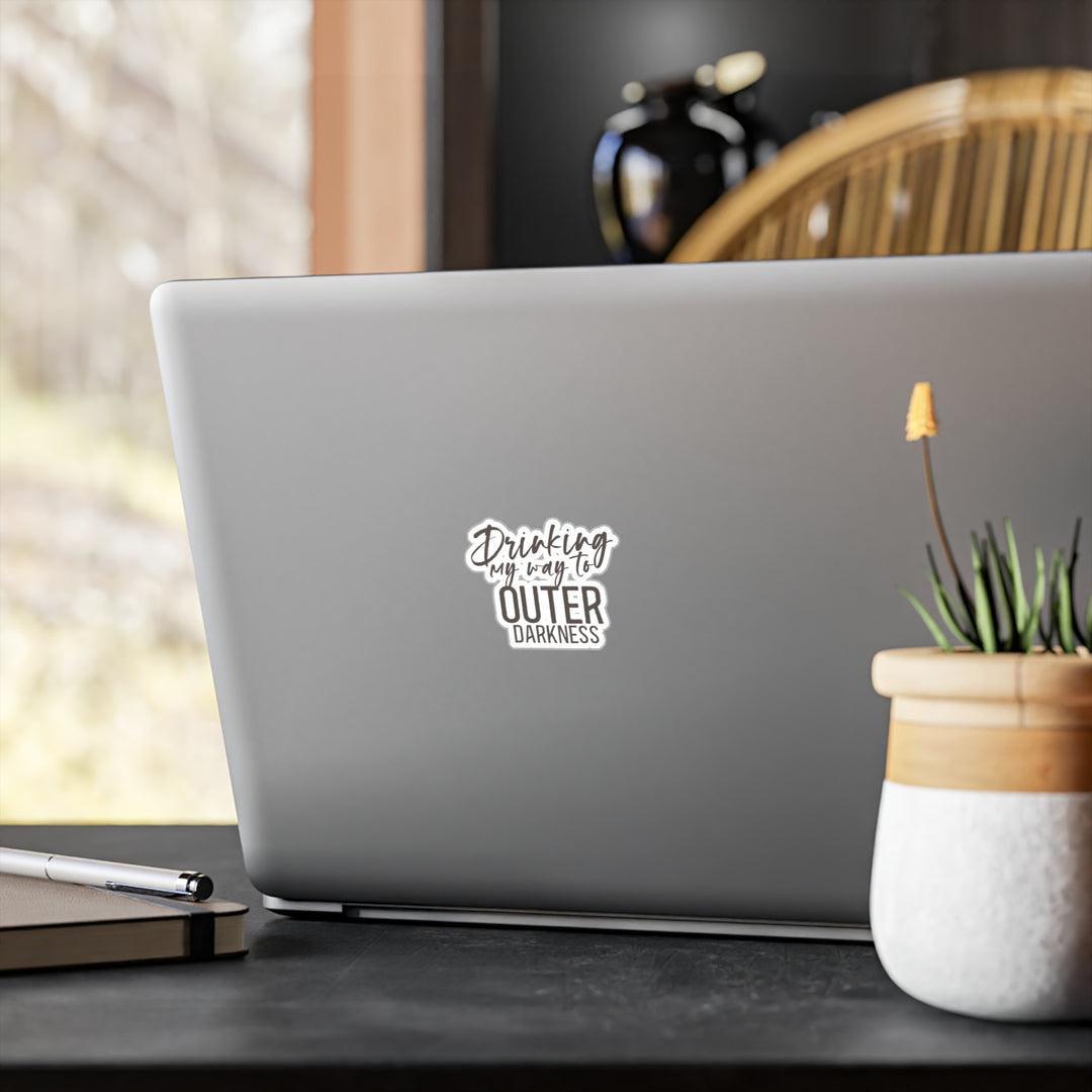 A white vinyl sticker with a black text and design, placed on a laptop, with a potted plant and stationery items in the background.